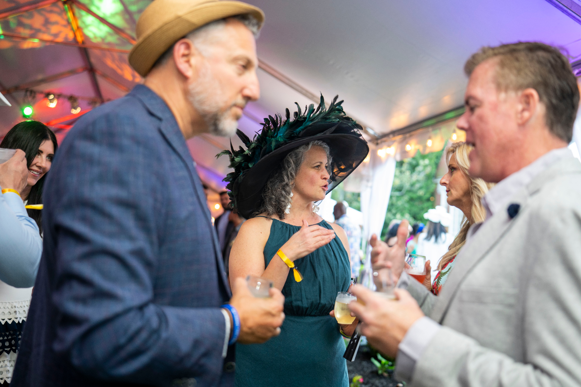 A guest in a feathered hat mid-conversation under amber tent light