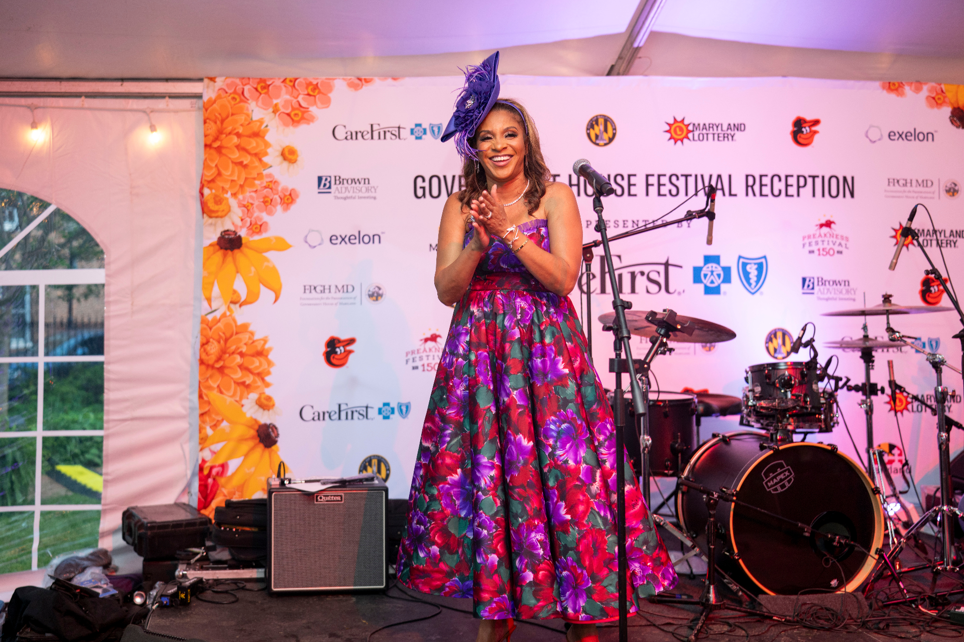 A speaker in a floral gown and purple fascinator on the Governor's House stage